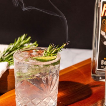 Gin and tonic with a spring of rosemary smoking sitting on a wood cutting board with a bowl of rosemary in the background.