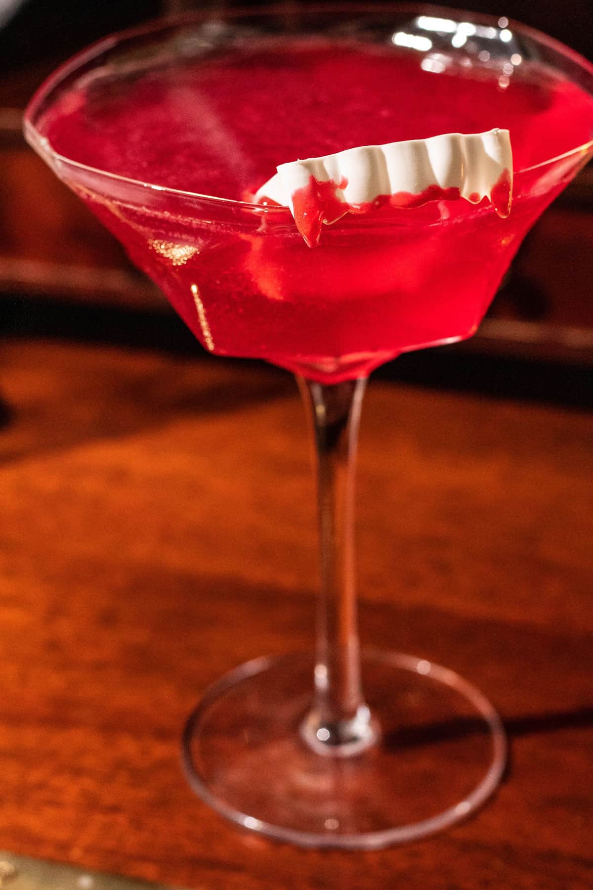 Close up of the vampire's kiss cocktail in a martini glass with a pair of toy vampire fangs with red icing on the teeth. The glass sits on a dark wood background.