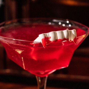 Top of the martini glass bowl with sparkling red cocktail and a pair of toy vampire fangs on the rim of the glass covered in red icing.