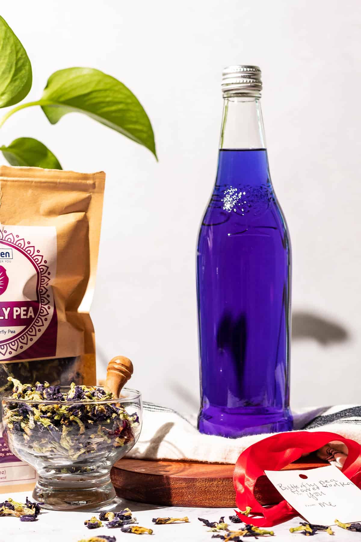 Glass bottle of purple butterfly pea infused vodka with a bag and bowl of the tea on the side and a green plant in the background. A red ribbon and tag are seen in the front.