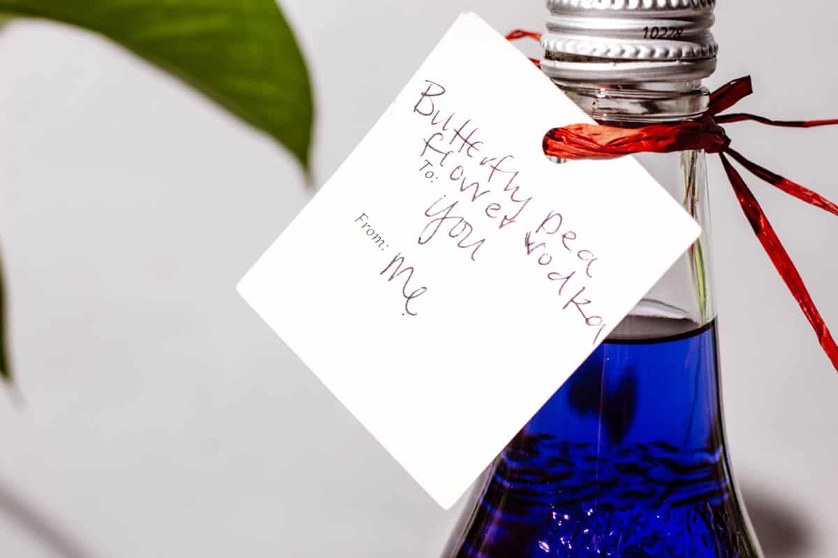 Close up of the purple vodka bottle with a gift tag that says "butterfly pea flower vodka, to: you, from: me" tied with a red string.