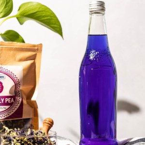 Glass bottle of purple butterfly pea infused vodka with a bag and bowl of the tea on the side and a green plant in the background. A red ribbon and tag are seen in the front.