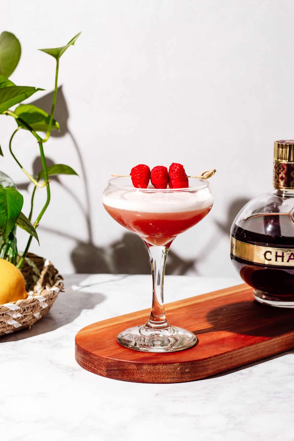 Coupe glass of chambord sour sitting on wood cutting board with a green plant and bottle of chambord in the background.