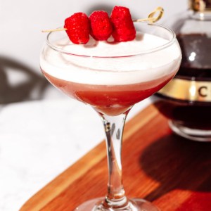 Coupe glass of chambord cocktail garnished with raspberries with a bottle of chambord in the background.