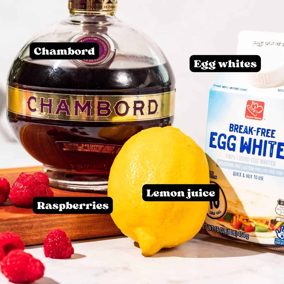 Ingredients shown on a wood cutting board including chambord raspberry liqueur, lemon, carton of egg whites, and fresh raspberries.