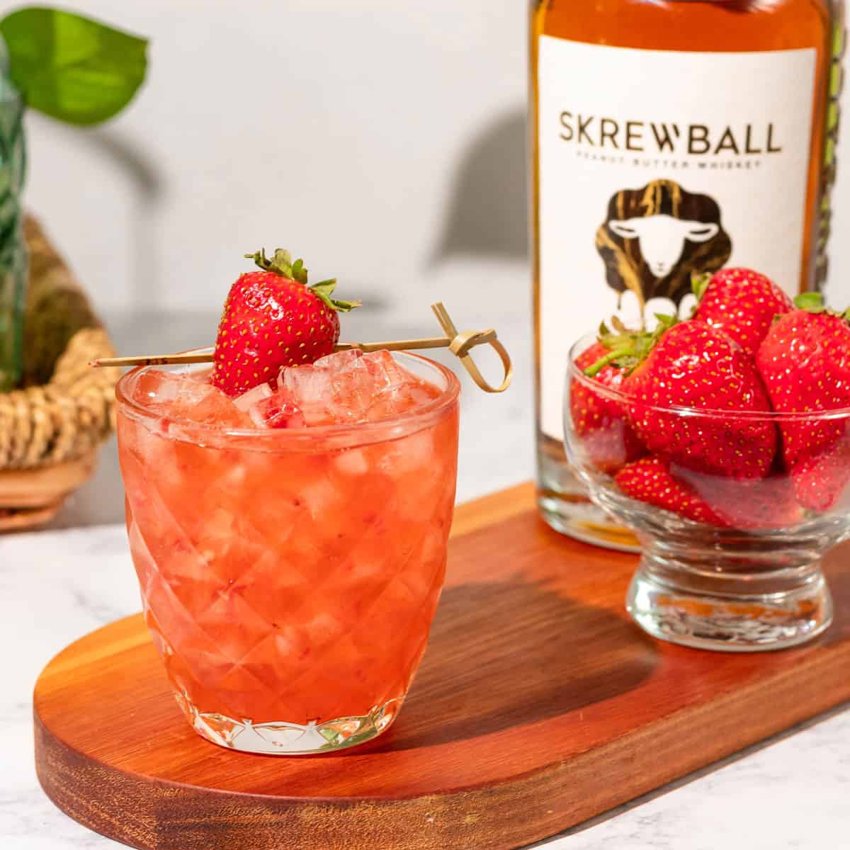Peanut butter and jelly cocktail sitting on a wood cutting board with a bowl of strawberries and a bottle of Skrewball peanut butter whiskey in the background.