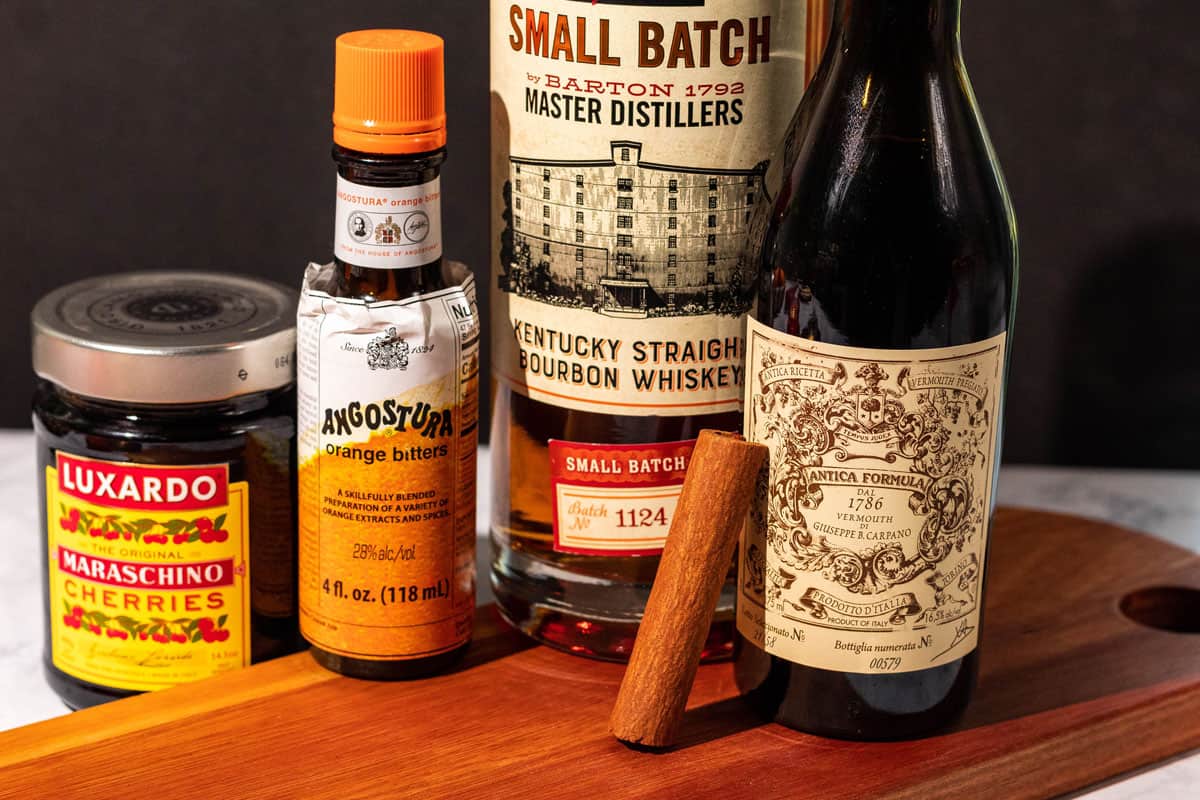 Ingredients for the smoked manhattan shown on a cutting board including bourbon, sweet vermouth, cinnamon stick, orange bitters, and luxardo cherries.