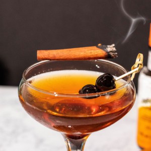 Close up of the smoked manhattan cocktail in a coupe glass garnished with cherries and a smoking cinnamon stick with a black and white background.