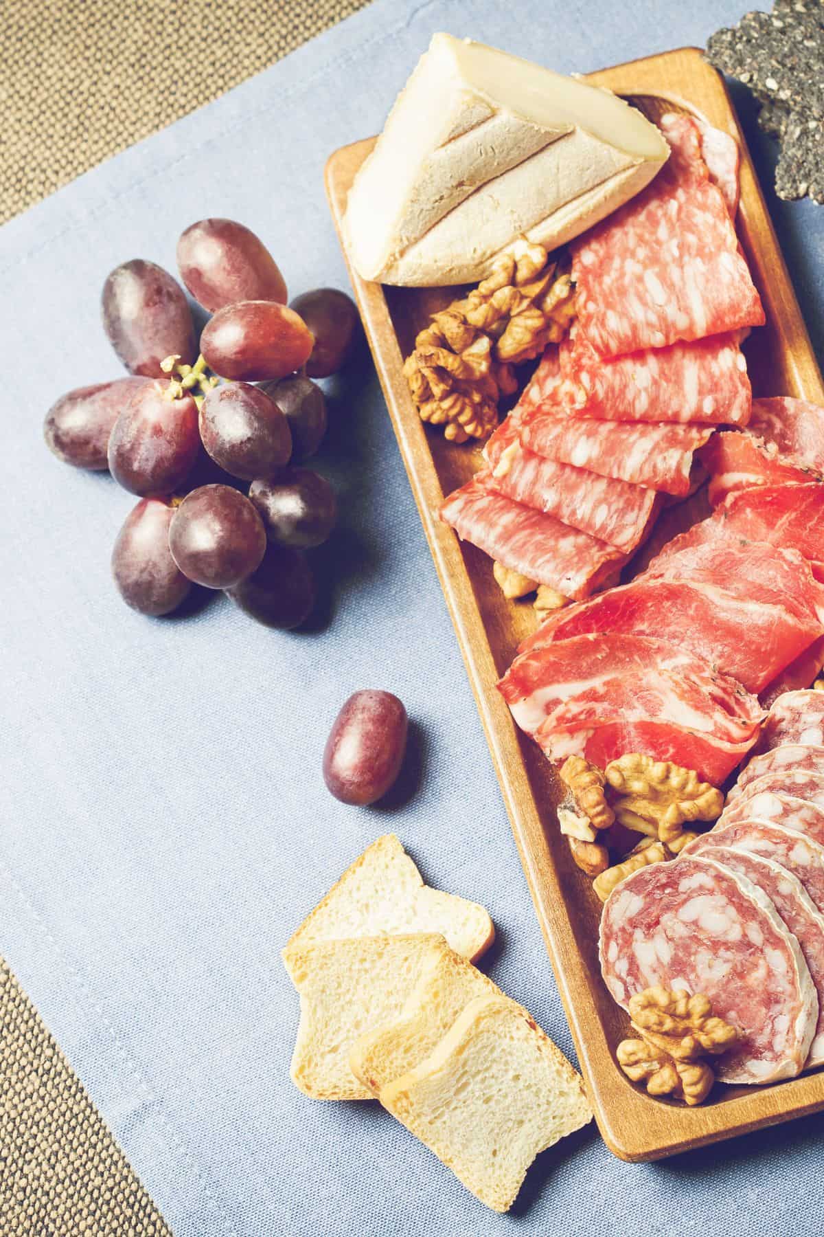 Different types of cured meats on a wood cutting board with grapes.