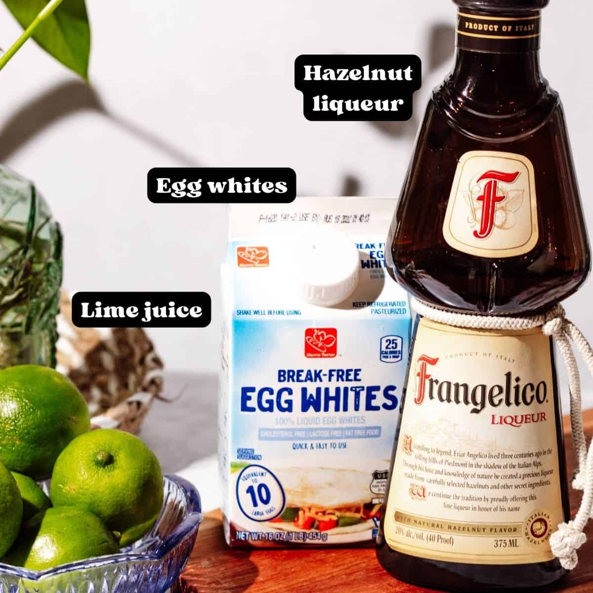 A bottle of frangelico, carton of egg whites and a bowl of limes with a green plant in the background. Text overlay says "hazelnut liqueur, egg whites, lime juice" in black and white lettering.