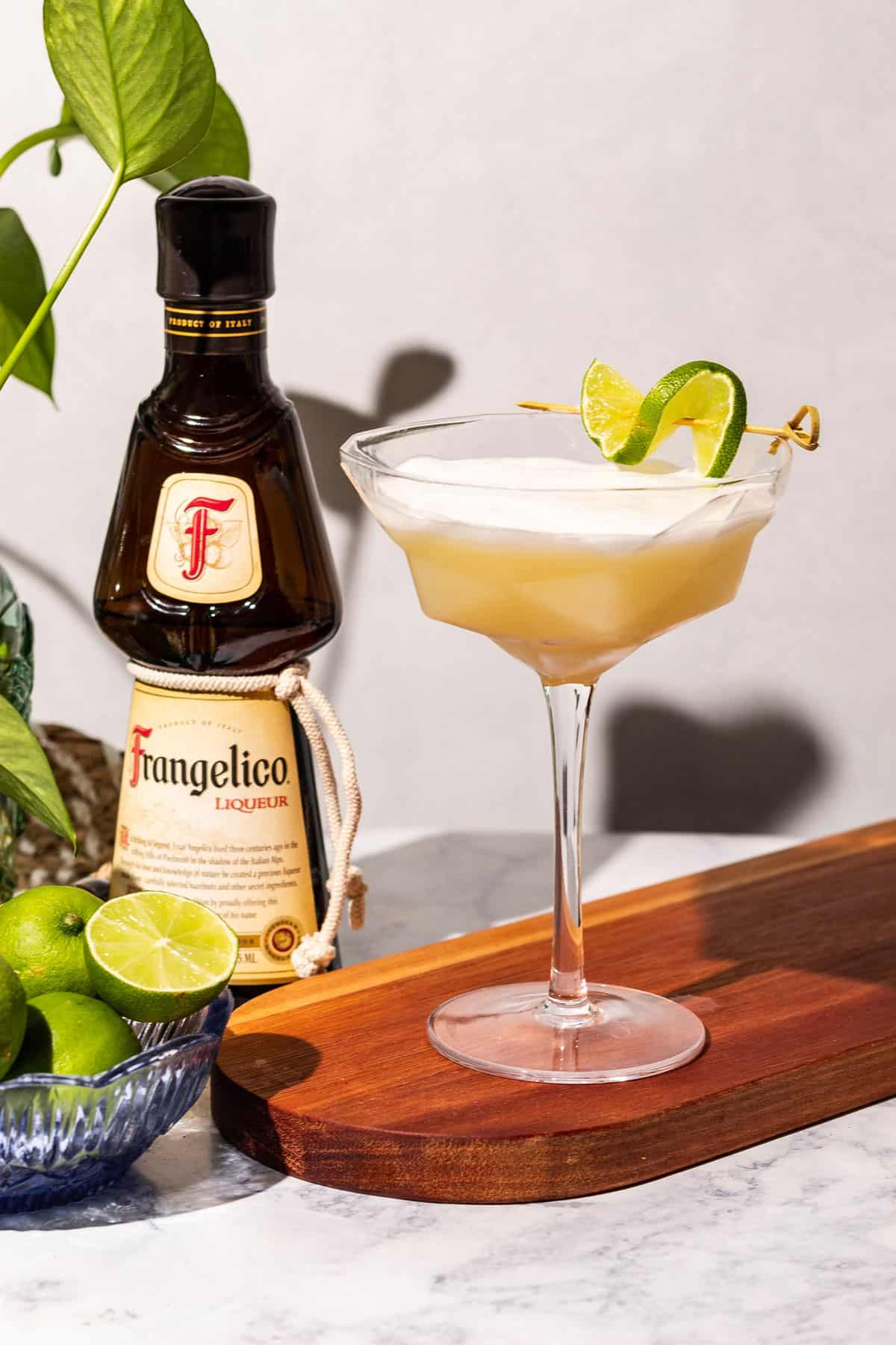 A couple glass of Frangelico Sour sits on a wood cutting board with limes, a green plant, and a bottle of frangelico sits in the background.