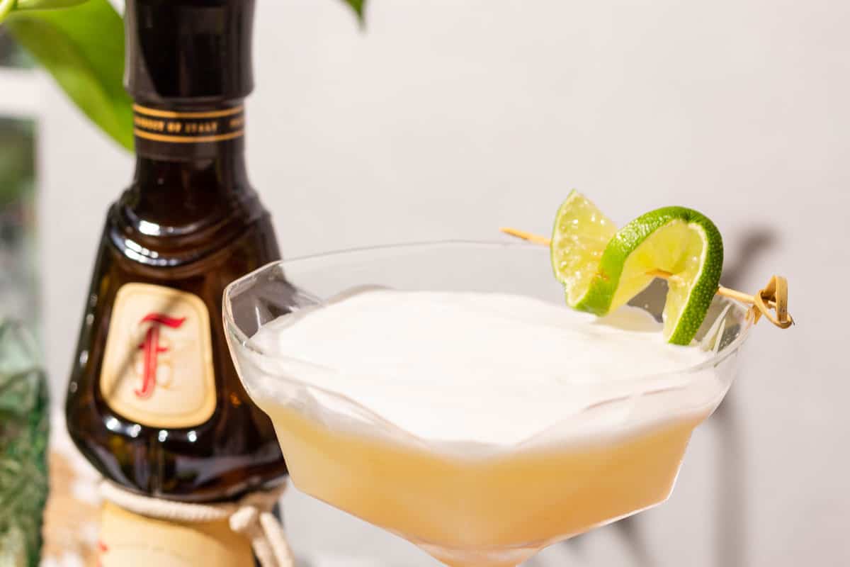 Close up of the top of the coupe glass showing the white foam and garnish of lime with a bottle of frangelico in the background.