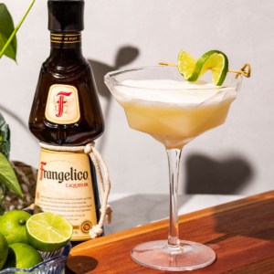 A couple glass of Frangelico Sour sits on a wood cutting board with limes, a green plant, and a bottle of frangelico sits in the background.
