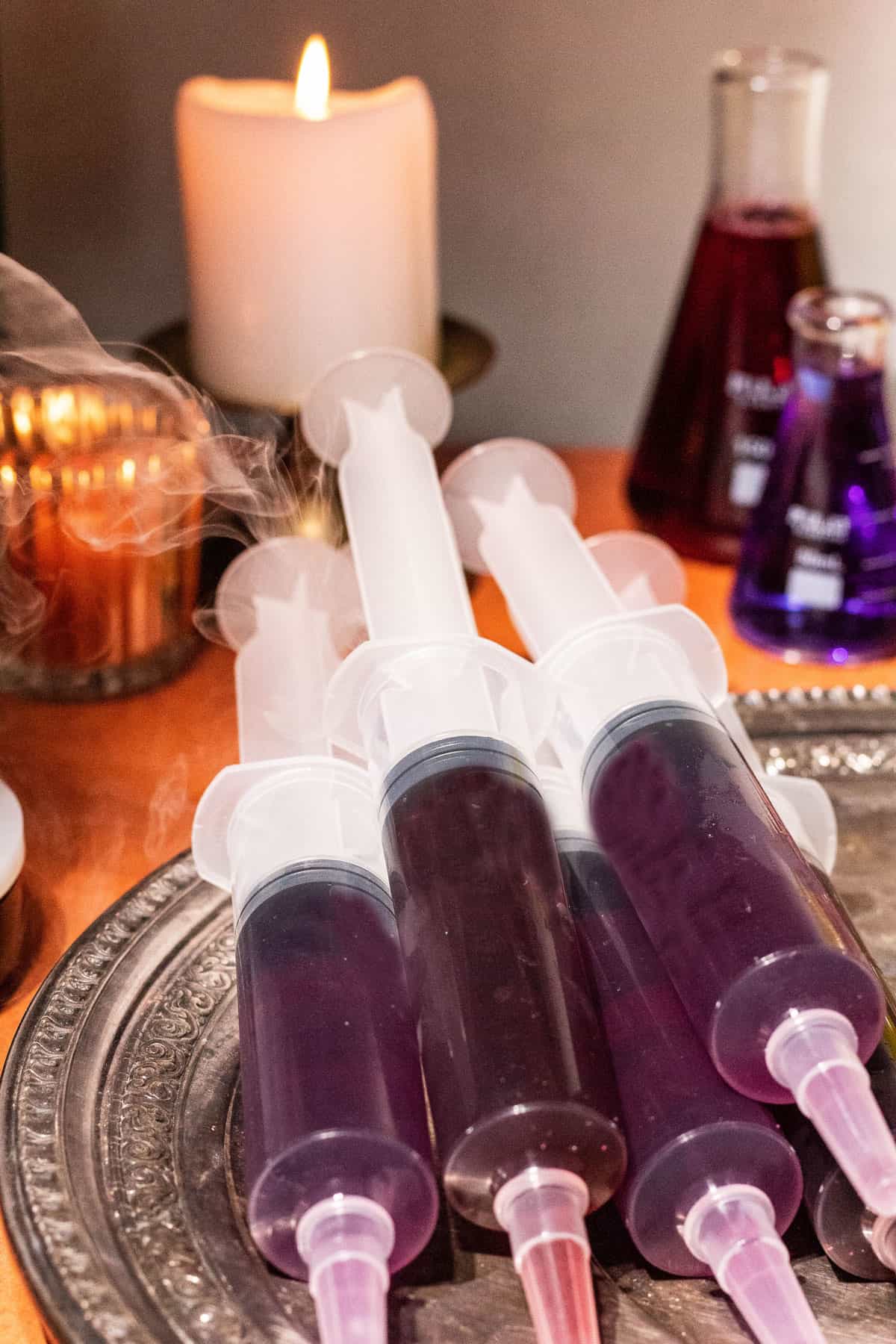 5 Halloween jello shot syringes stacked on a silver platter with a candle and fog in the background. 