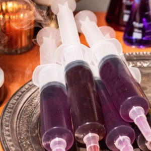 5 Halloween jello shot syringes stacked on a silver platter with a candle and fog in the background.