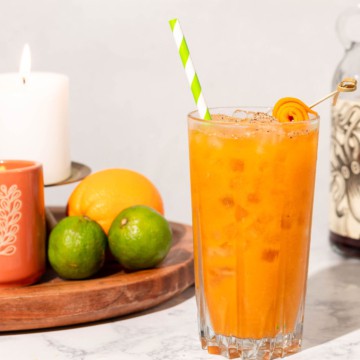 Tall glass of pumpkin cocktail with green and white striped straw and a wood board full of citrus and candles in the background.