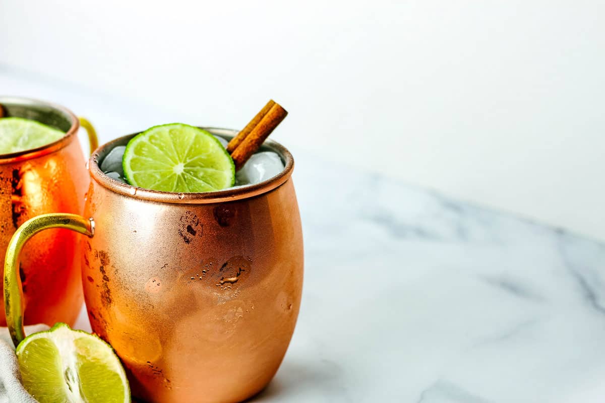 Copper mug garnished with lime and cinnamon stick on a white counter.
