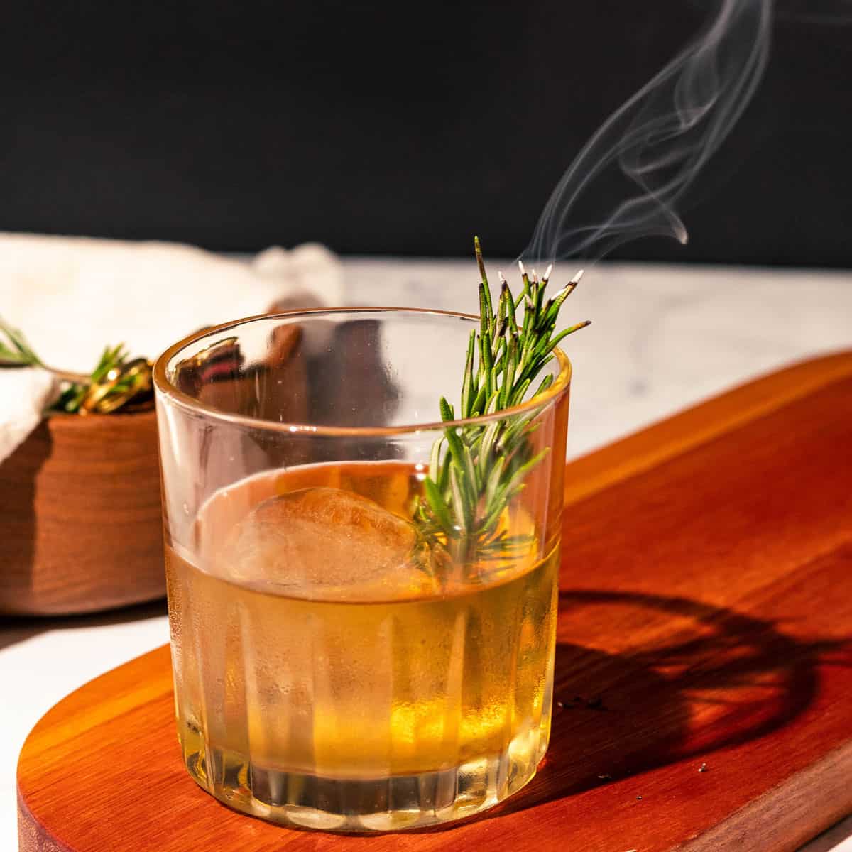 Old fashioned on a cutting board with a smoking rosemary sprig.