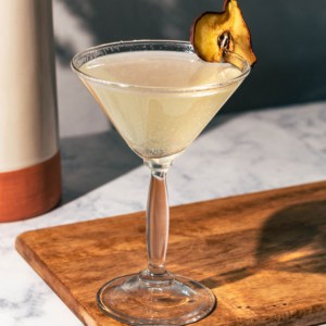 Cocktail on a wood board with dried flower vase in background.