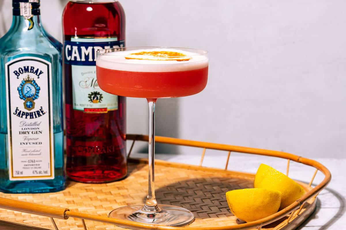 Negroni sour in a long stem coupe glass with bottles of Campari and gin in the background.