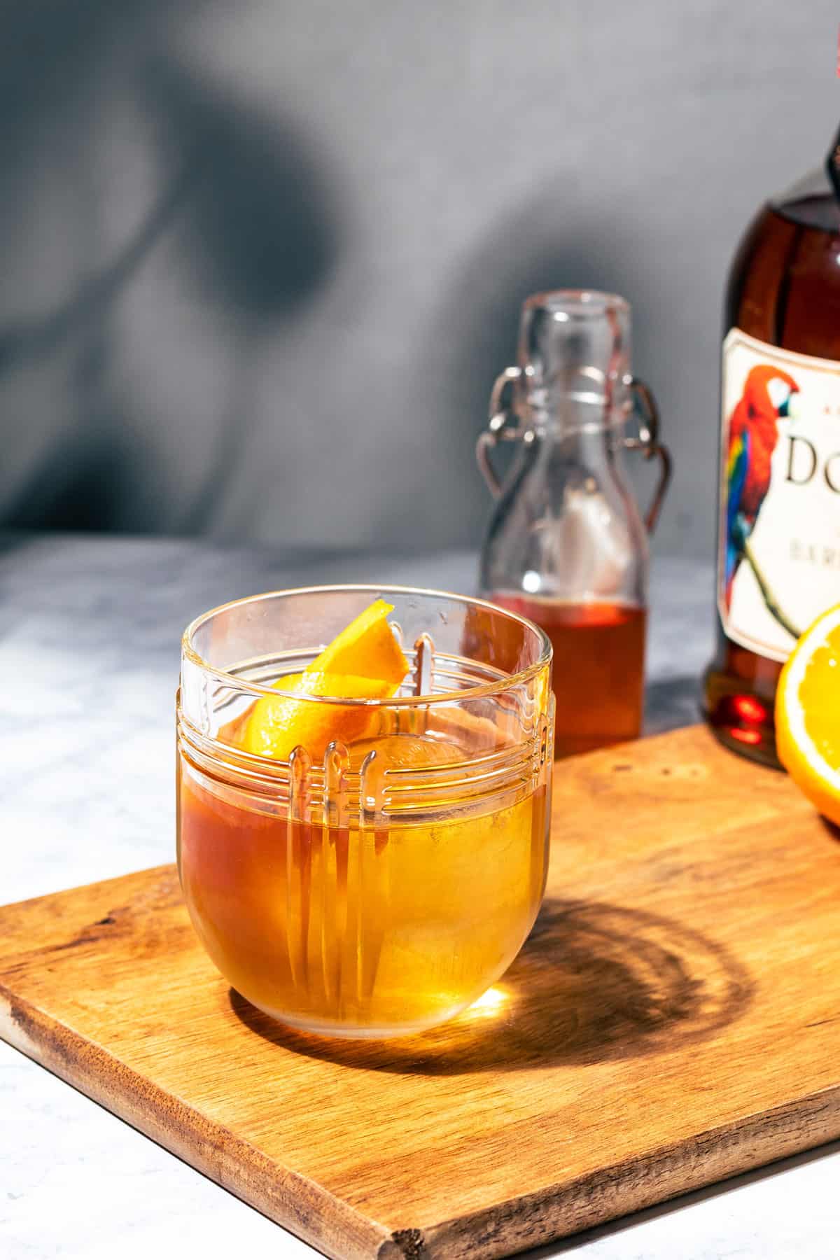 Old fashioned sitting on a wood cutting board with a bottle of rum, syrup, and citrus in the background.
