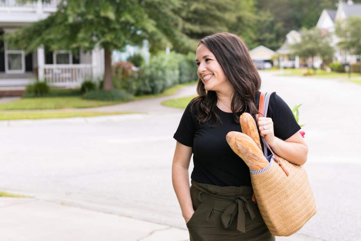 Melissa in black shirt, green pants, smiling to her right while carrying a straw bag with bread and flowers inside.