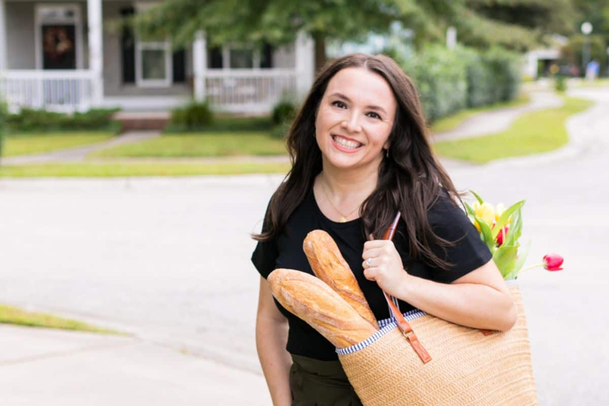 Melissa in black shirt, green pants, facing forward, while carrying a straw bag with bread and flowers inside.