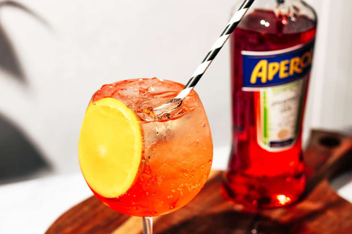 Aperol spritz with a black and white straw and a bottle of Aperol in the background.