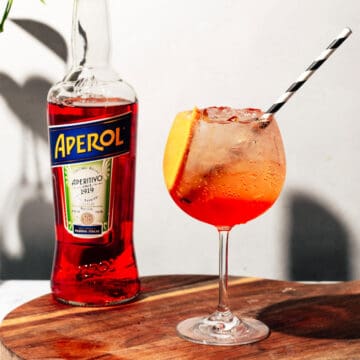 Aperol spritz sitting on a wood cutting board with a bottle of Aperol in the background.