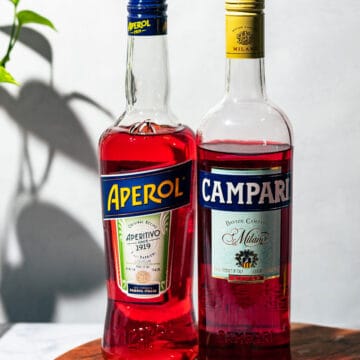 A bottle of Aperol and Campari sitting on a wooden cutting board with a green plant in the background.
