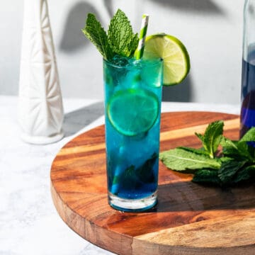 Blue mojito sitting on a round cutting board with mint and a bottle of blue curacao on the side.