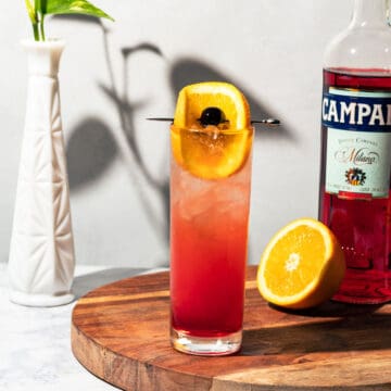 Campari soda cocktail on a round wood cutting board with a bottle of Campari and an orange half in the background.