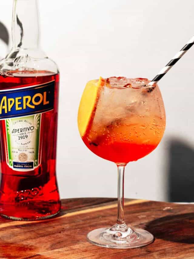 Aperol spritz sitting on a wood cutting board with a bottle of Aperol in the background.