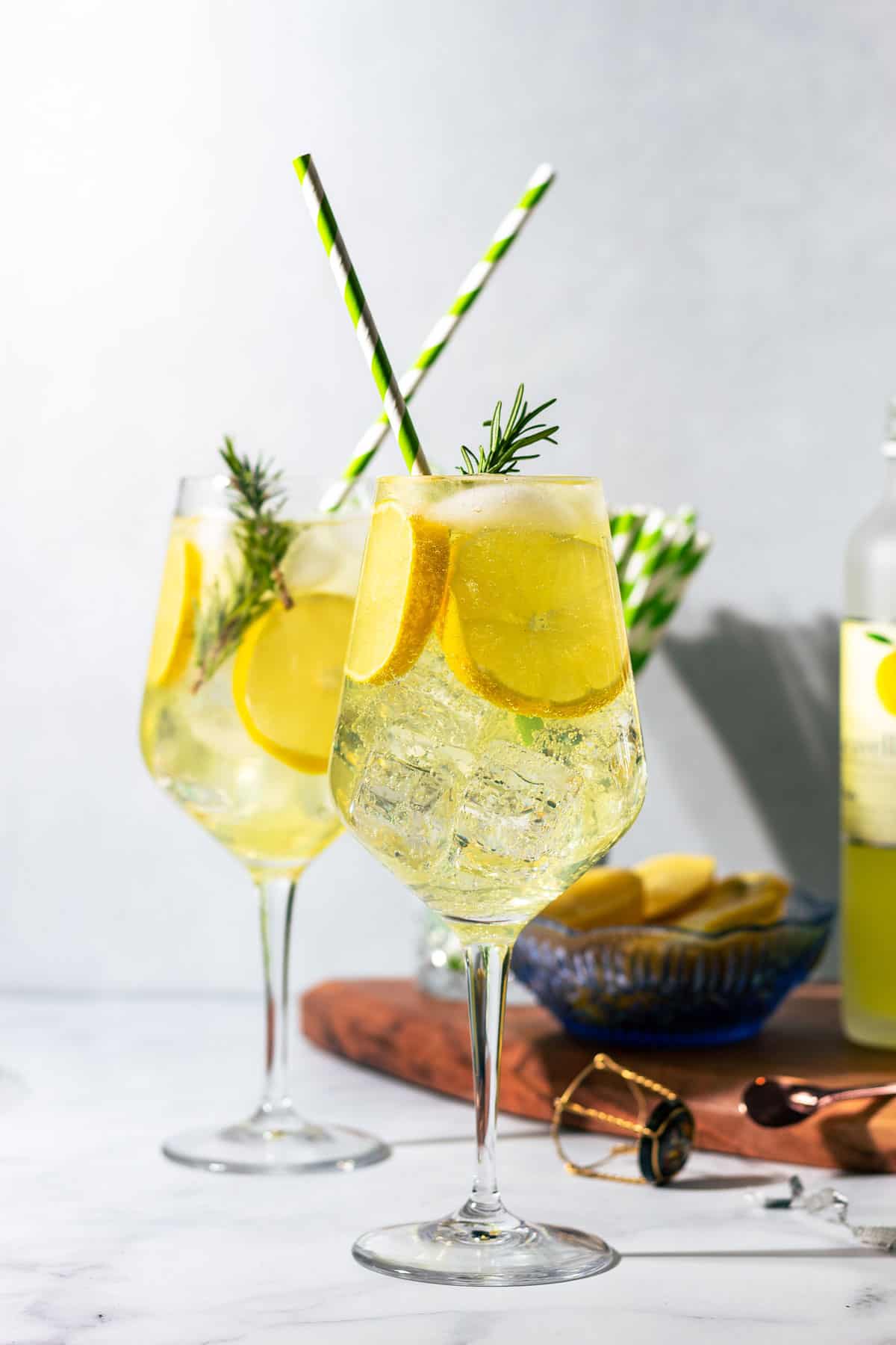 Two wine glasses of Limoncello Spritz with green striped straws on a white countertop.