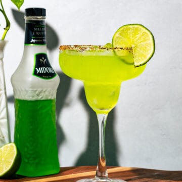 Midori Margarita on a wood cutting board with a bottle of midori, lime half, and a plant in the background.
