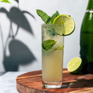 Mojito on a round wooden cutting board with a bottle of sparkling wine and a lime half in the background.