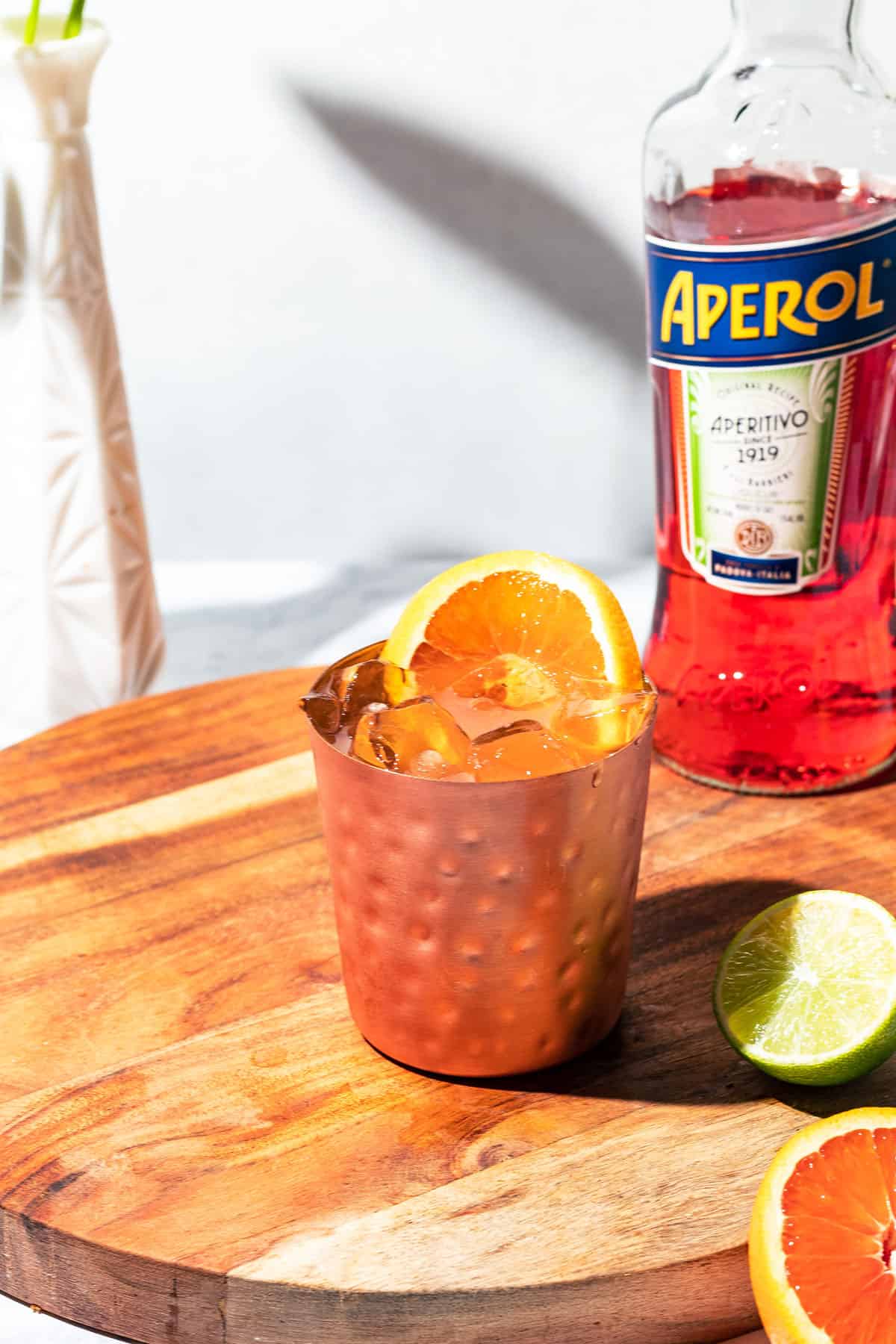 Mule cocktail on a wooden surface with a bottle of Aperol and citrus fruit on the side.