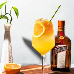 Spritz in a wine glass with a bottle of Cointreau, orange half, and green plant in the background.