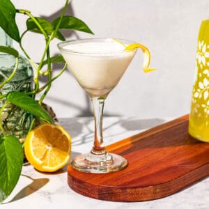 Cocktail in a martini glass with a green plant, lemon half, and bottle of limoncello in the background.