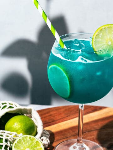 Large wine glass with the Mermaid Water Margarita with a netted bag with limes next to it.