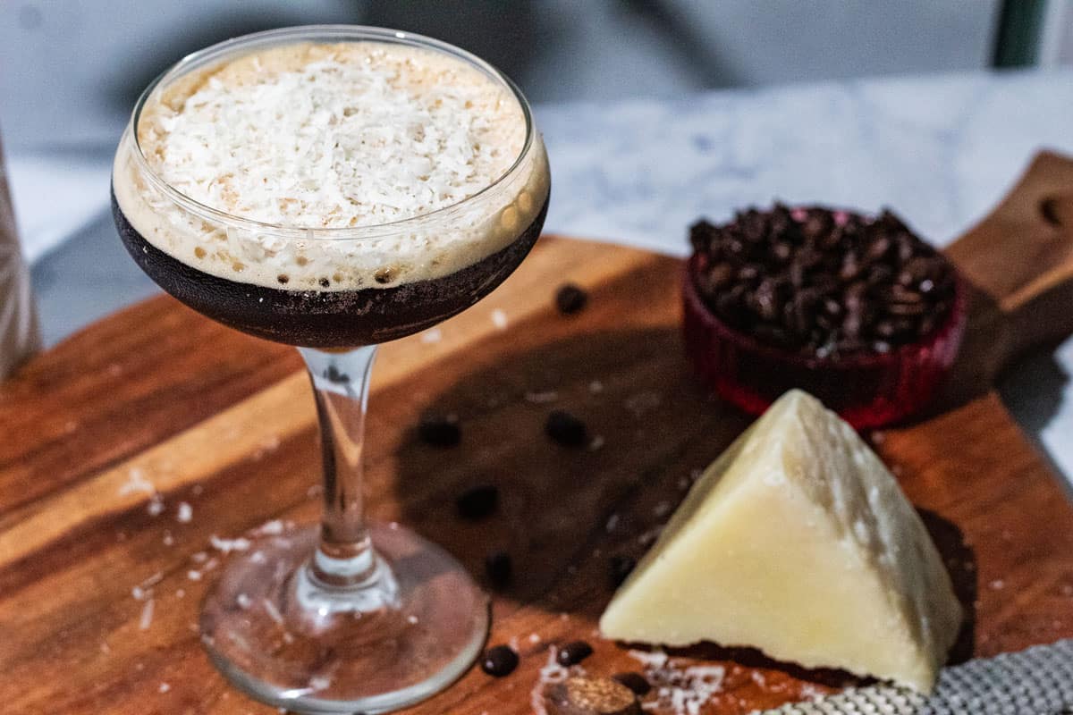 Espresso martini on a wood surface with a wedge of parmesan cheese, whole nutmeg, and a bowl of coffee beans in the background.