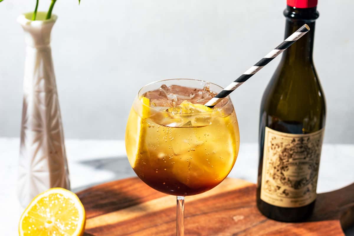 Spritz with a black and white straw on wood surface with a bottle of vermouth and lemon half on the side.