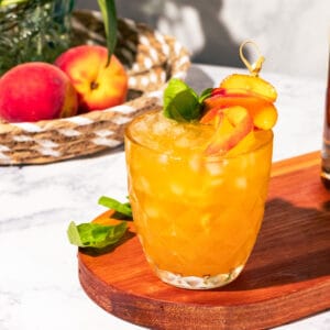 Bourbon peach smash on a wood cutting board with peaches in a basket in the background.