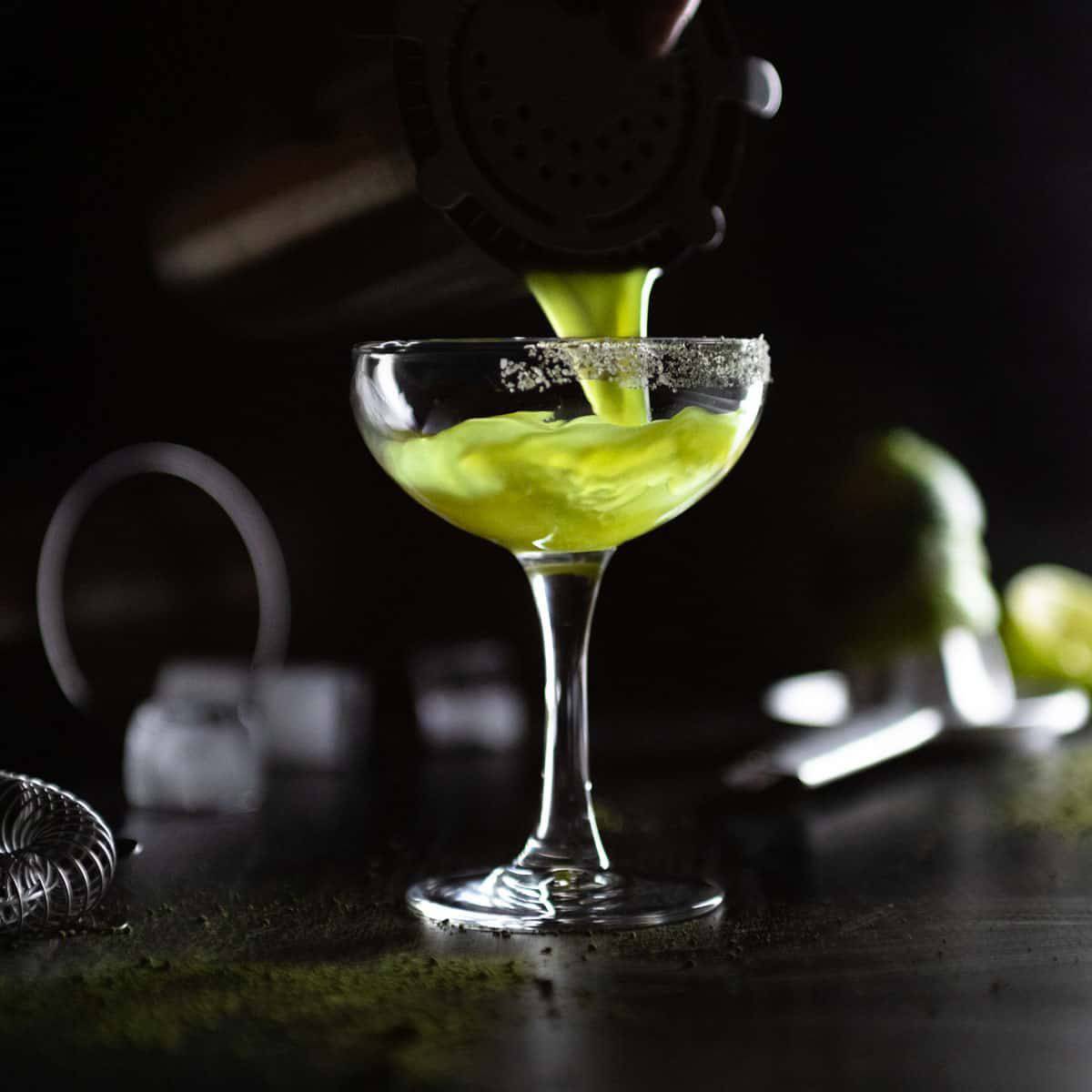 Green cocktail being poured into a coupe glass.