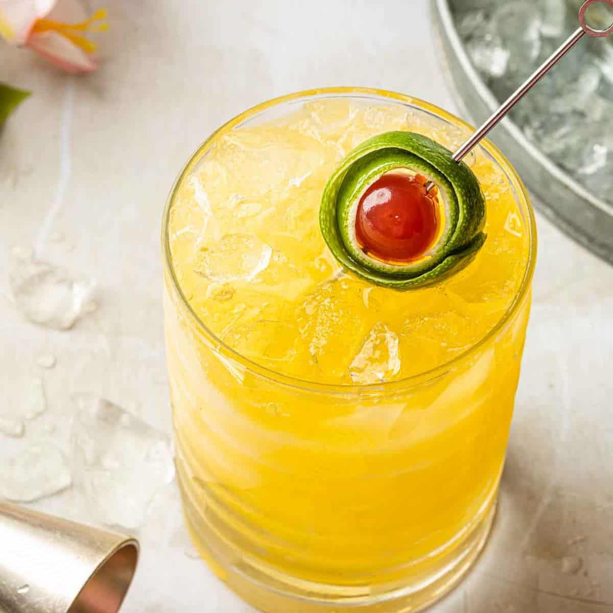 Yellow cocktail garnished with a lime peel wrapped around a cocktail cherry.