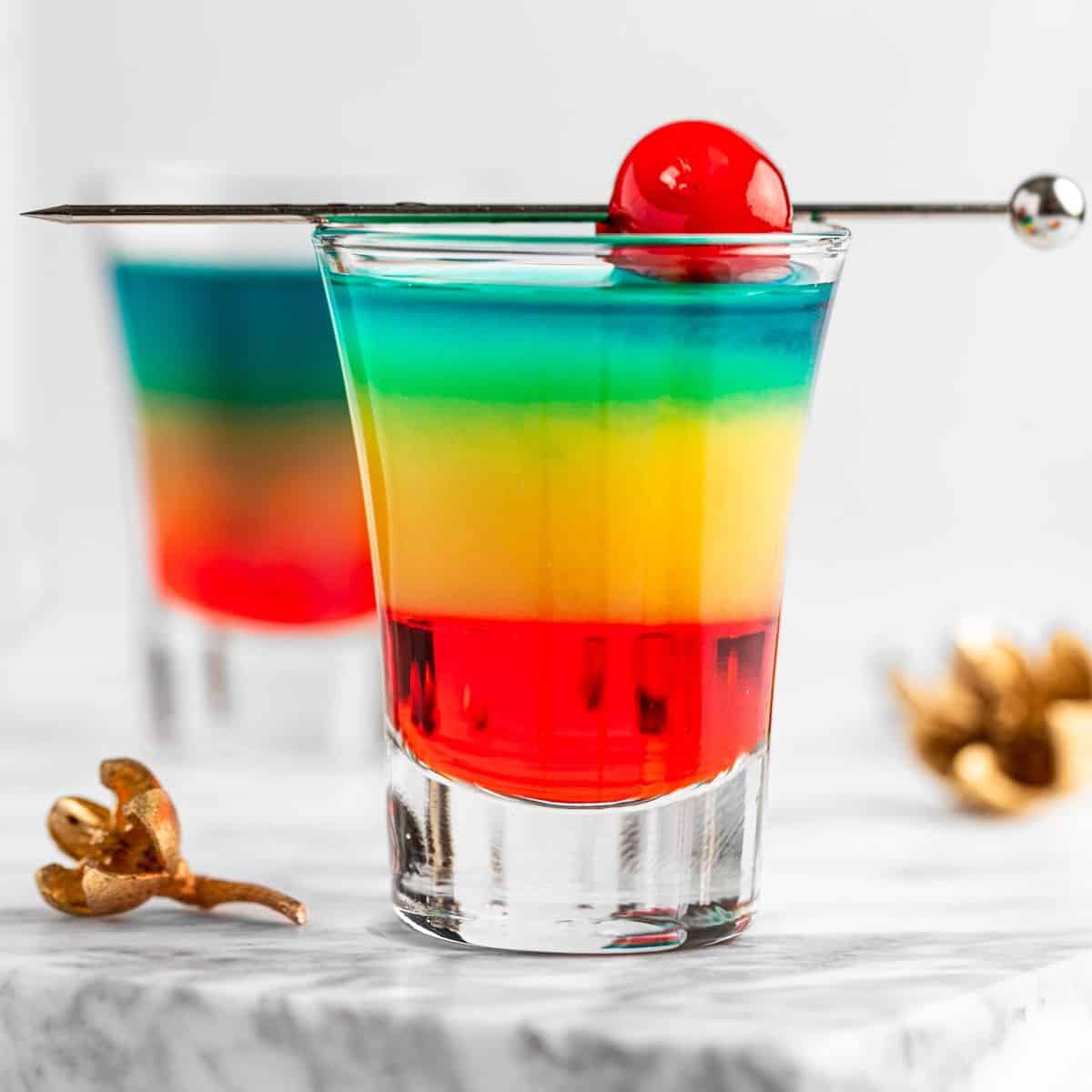 Layered rainbow drink in a shot glass garnished with a cherry.