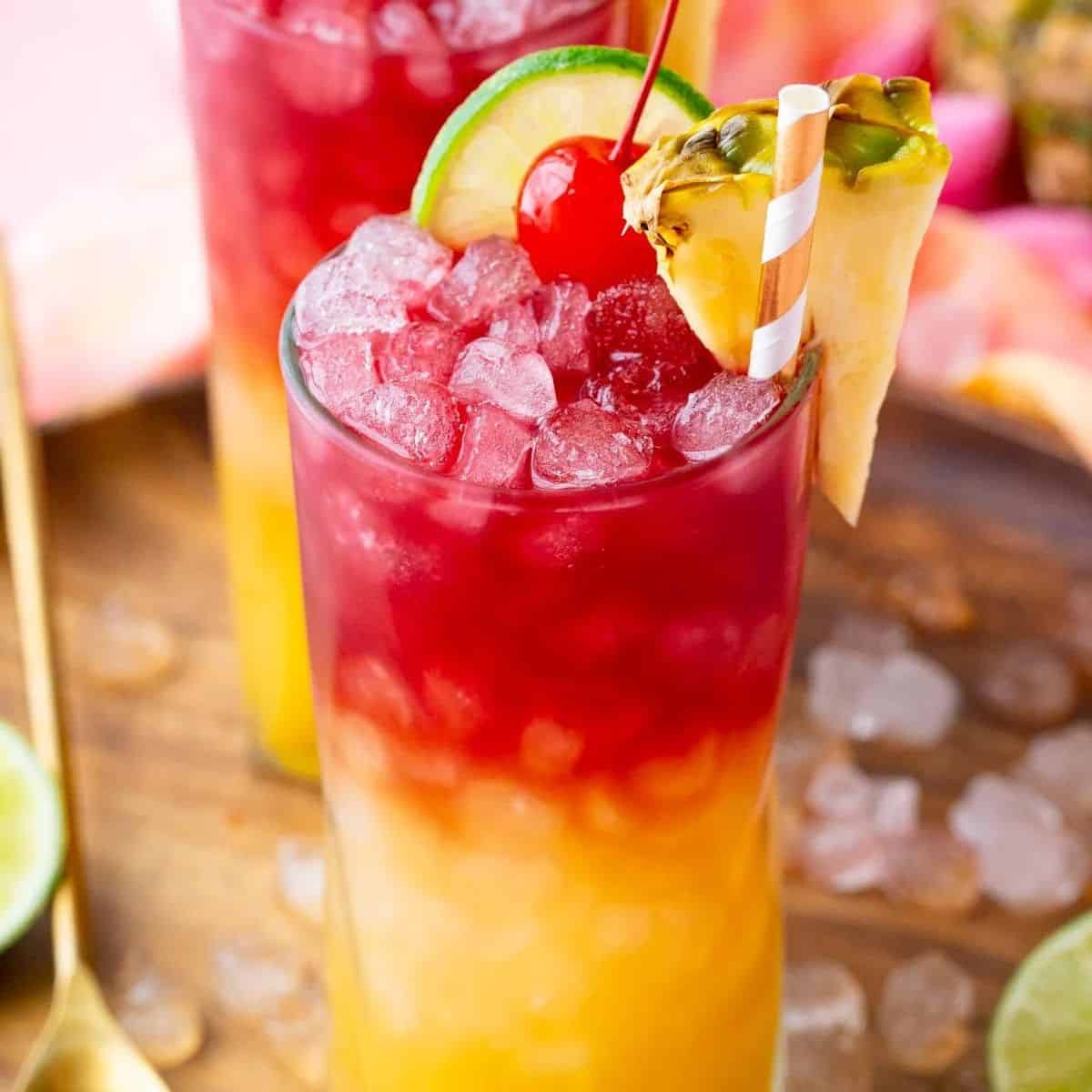 Yellow and red cocktail garnished with a cherry, lime slice, and pineapple slice.
