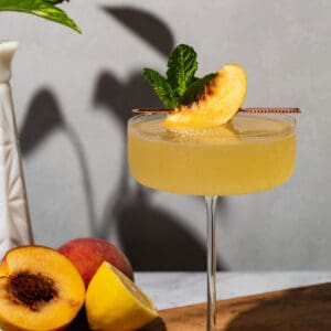 The cocktail in a coupe glass with peach and lemon halves in the background.