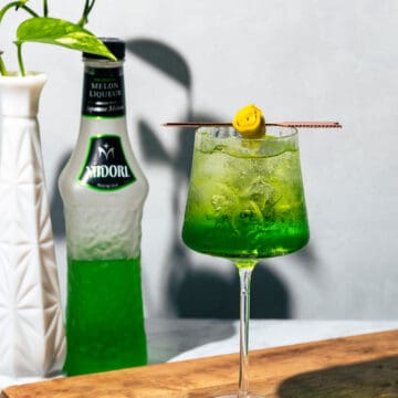 Green spritz cocktail on a wood surface garnished with a rolled up lemon twist on a cocktail pick.