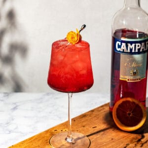 Campari tonic on a wood surface with a bottle of Campari and orange half in the background.
